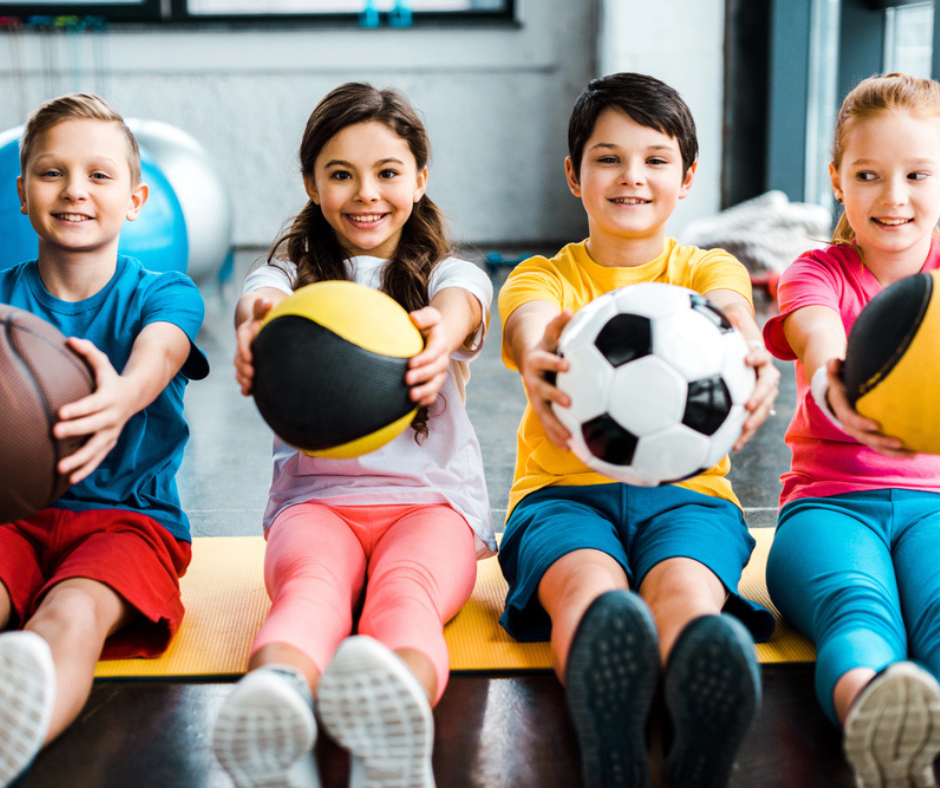 girls and boys sit on the floor of a school gym and hold out various types of sports balls for soccer, basketball, and volleyball